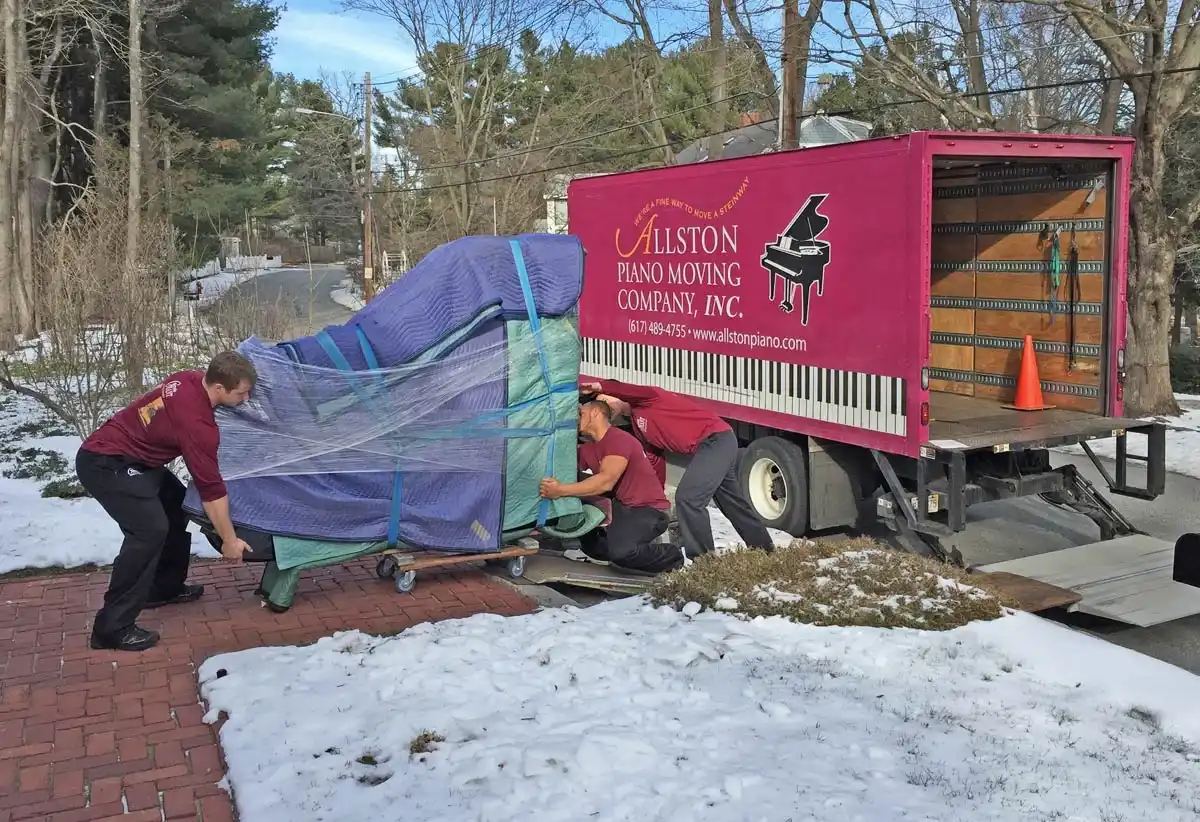 Movers wheeling a blanket-wrapped piano to a piano moving truck in the snow