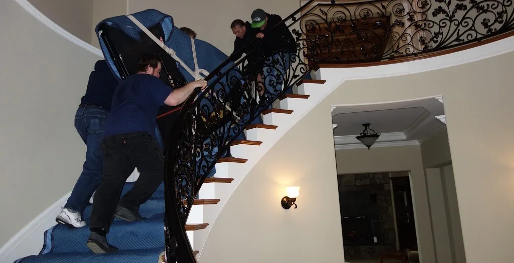 Movers carrying a wrapped grand piano up a curved staircase